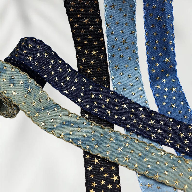 May include: Assortment of denim ribbons in various shades of blue and black, adorned with gold star patterns. The ribbons have a frayed edge and are arranged in a diagonal composition, showcasing their texture and design.
