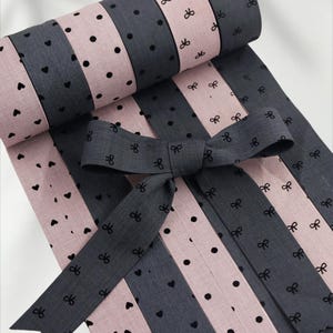 Op de afbeelding: Een opgerolde stof met afwisselende roze en grijze strepen. De roze strepen hebben zwarte hartjes, terwijl de grijze strepen zwarte stippen en strikpatronen hebben. Een grijze strik is bovenop geplaatst, met strikpatronen.