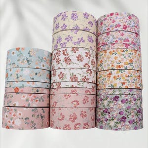 Könnte beinhalten: Eine Sammlung von Washi-Tape-Rollen mit floralen Mustern in verschiedenen Farben. Die Rollen sind gestapelt und zeigen Designs mit rosa, lila und blauen Blumen auf hellem Hintergrund. Das Klebeband wird wahrscheinlich für Bastelarbeiten und dekorative Zwecke verwendet.