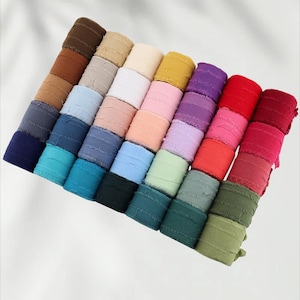 Könnte beinhalten: Eine Sammlung farbenfroher Stoffrollen in verschiedenen Farbtönen, darunter Braun, Beige, Blau, Grün, Rosa, Lila und Rot. Die Rollen sind diagonal angeordnet und zeigen die vielfältige Farbpalette und das strukturierte Material. Der Stoff scheint ein strukturierter Gaze oder Leinen zu sein.