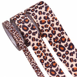 Peut inclure: Trois rouleaux de ruban gros-grain avec un motif léopard répétitif. Le design se compose de taches brun foncé et orange sur un fond beige clair. Les rubans en tissu texturé sont présentés dans des largeurs différentes.