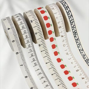 May include: A collection of white measuring tapes with black markings, including inch and centimeter scales. One tape features red apple patterns, and another displays the alphabet. The tapes are rolled.