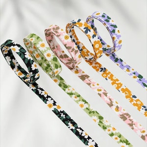 Könnte beinhalten: Eine Auswahl an floral gemusterten Bändern in verschiedenen Farben, darunter Schwarz, Grün, Rosa, Orange und Lila. Jedes Band zeigt ein sich wiederholendes Gänseblümchen-Design mit weißen Blütenblättern und gelben Zentren. Die Bänder sind diagonal angeordnet.