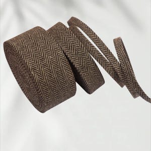 Könnte beinhalten: Eine Rolle braun-beiges Fischgrätenband. Das Band ist eng aufgerollt, mit mehreren Strängen, die sich entwirren. Der Stoff hat eine gewebte Textur und die Farbe ist ein warmer, erdiger Ton. Ideal für Bastelarbeiten.