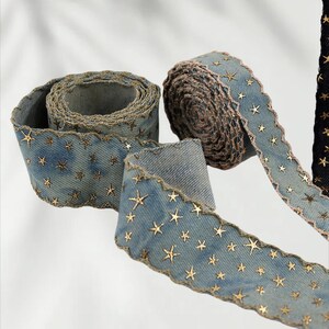 May include: Denim ribbon with gold star embellishments. The ribbon has a scalloped edge and is rolled into several spools. The denim is a light blue color, and the stars are a metallic gold. The ribbon is suitable for crafts and decorations.