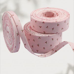 Könnte beinhalten: Drei Rollen rosa-weiß gestreiftes Band mit Kirschmuster. Eine Rolle ist abgerollt und zeigt den Rand des Bandes. Das Band eignet sich für Bastelarbeiten, Nähen und Geschenkverpackungen. Das Band ist aus Stoff.
