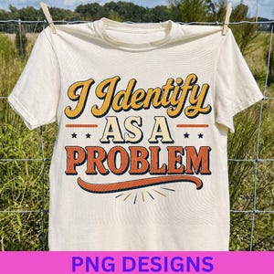 May include: A cream-colored short-sleeve t-shirt featuring the text I Identify AS A PROBLEM in a retro-style font. The words are printed in yellow, cream, and orange with dark blue outlines, accented by small stars and a curved orange line.