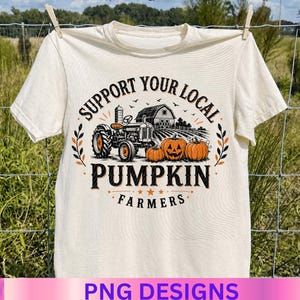 May include: A cream-colored t-shirt featuring a graphic print of a tractor in a field with three pumpkins and a barn. The text reads "SUPPORT YOUR LOCAL PUMPKIN FARMERS" in black lettering, with orange accents and small stars surrounding the design.