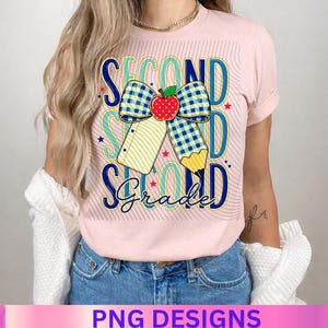 May include: A light pink t-shirt featuring a graphic design with the text SECOND GRADE in blue and teal. The design includes a blue and white checkered bow, a red apple, a yellow pencil, and small red and blue stars on a background of thin lines.