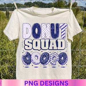 May include: Cream-colored t-shirt with the words "DONUT SQUAD" in blue and white. Below the text are five blue donuts with different designs. The shirt is hanging on a clothesline with a green and brown background.