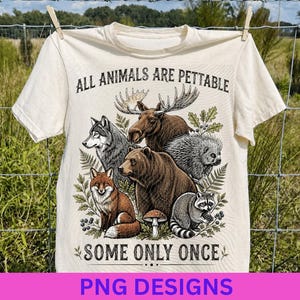 May include: A cream-colored short-sleeve t-shirt featuring a graphic illustration of a moose, wolf, bear, porcupine, fox, and raccoon surrounded by foliage. The text reads ALL ANIMALS ARE PETTABLE SOME ONLY ONCE in a dark brown serif font.