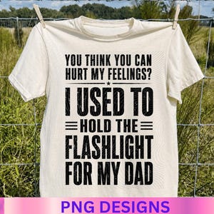 Może przedstawiać: Kremowa koszulka z czarnym napisem "YOU THINK YOU CAN HURT MY FEELINGS? I USED TO =HOLD THE= FLASHLIGHT FOR MY DAD". Koszulka wisi na sznurku do prania z zielono-brązowym tłem.