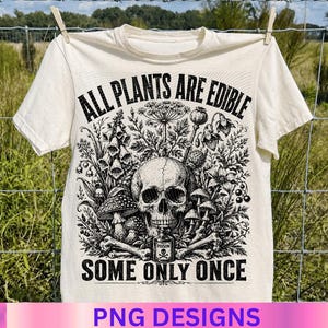 May include: A cream-colored t-shirt featuring a black graphic print of a human skull surrounded by various plants, mushrooms, and bones. Below the skull is a small bottle labeled POISON. The text reads ALL PLANTS ARE EDIBLE SOME ONLY ONCE.
