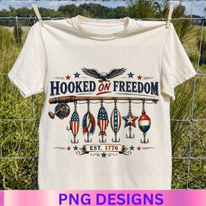 May include: A cream-colored t-shirt featuring a graphic of a fishing rod with various patriotic lures hanging from it. Above the rod is an eagle and the text HOOKED ON FREEDOM. Below the lures is a banner with the text EST. 1776 and small stars.