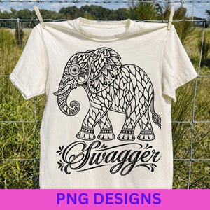 May include: A cream-colored short-sleeve t-shirt featuring a large black graphic of an elephant decorated with intricate floral and geometric patterns. Below the elephant, the word "Swagger" is printed in a stylized, cursive black script font.