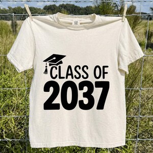 Può includere: T-shirt color crema con la scritta "CLASS OF 2037" in grassetto nero. Sopra il testo, una grafica di cappello di laurea. La maglietta è appesa a un filo per stendere i panni con uno sfondo naturale.