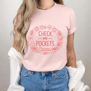 Może przedstawiać: Jasnoróżowa koszulka z czerwonym napisem "CHECK MY POCKETS", otoczonym wieńcem z liści, owadów i innych ilustracji o tematyce przyrodniczej. Koszulka wykonana jest z miękkiego materiału.