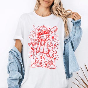May include: White t-shirt featuring a red cartoon monkey wearing sunglasses, a jacket, and holding a baseball bat. The monkey is smiling with a gold chain around its neck. Red paint splatter design around the monkey.