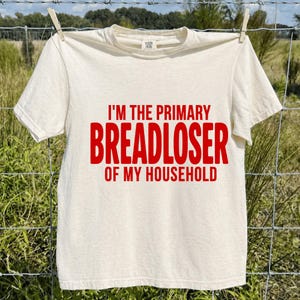 Könnte beinhalten: Cremefarbenes T-Shirt mit dem Text "I'M THE PRIMARY BREADLOSER OF MY HOUSEHOLD" in roten Blockbuchstaben. Das T-Shirt hängt an einer Wäscheleine mit einer Wäscheklammer.