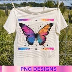 May include: A cream-colored t-shirt featuring a large, vibrant butterfly graphic with a gradient of purple, pink, and orange. The design includes thin black wavy lines, small star icons, and horizontal bands with watercolor-style paint splatters.