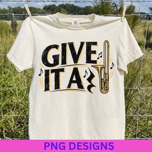 Könnte beinhalten: Ein cremefarbenes Kurzarm-T-Shirt mit dem schwarzen Schriftzug GIVE IT A, bei dem eine goldfarbene Posaune den Buchstaben L ersetzt. Das Design enthält kleine schwarze Musiknoten und eine abgenutzte Textur auf der Schrift und der Instrumentengrafik.