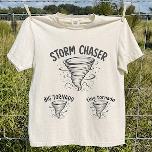 May include: Cream-colored t-shirt with a graphic of a tornado and the words "STORM CHASER." Below the main graphic are illustrations of two smaller tornadoes with the text "BIG TORNADO" and "tiny tornado."