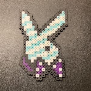 May include: A pixel art design of a stylized bunny head, crafted from colorful plastic beads. The design features a gray outline, with sections of white, light blue, and purple. The bunny's ears are upright, and the overall shape is symmetrical.