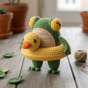 May include: A green crocheted frog toy with a beige belly and yellow eyes, wearing a yellow crocheted duck-shaped swim ring. The frog has small arms and legs, and the duck swim ring features a small orange beak and black eyes.