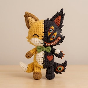 May include: A split-design crochet plush toy featuring a golden-yellow side with a winking eye and a black side with two eyes and a wide, toothy grin. The creature wears a green bow tie, has a red heart on its chest, and a tail with an eye.