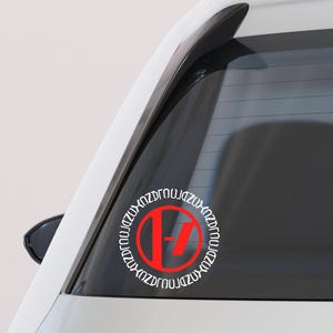 May include: A white car with a red and white circular sticker on the rear window. The sticker features a red "17" inside a white circle with repeating text around the edge. The car's taillight is visible.