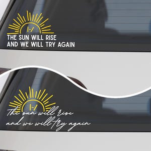 May include: A yellow and white vinyl decal for a car window featuring a stylised sun graphic with the text "THE SUN WILL RISE AND WE WILL TRY AGAIN" in a sans-serif font and a second version with the same text in a cursive script font.