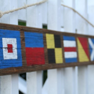 May include: A rustic wooden sign featuring nautical signal flags painted in blue, red, yellow, black, and white. The sign is suspended by a rope and is mounted on a white picket fence. The wooden sign is a decorative item.
