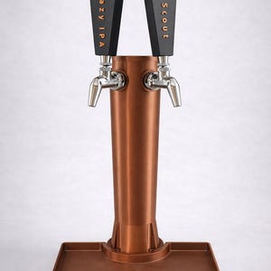 May include: A copper-colored beer tap with two spouts. The tap handles are black with the words "Hazy IPA" and "Scout" in gold lettering. The tap is mounted on a copper-colored base with a drip tray.