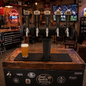May include: A beer tap system with five taps, labeled Pilsner, Hazy IPA, Stout, Lager, and Pale Ale, sits atop a wooden cooler. A glass of beer is on the cooler. The cooler has the text "Grand Beach Brewing" and "YETI" on it. A neon sign reads "Garage Beer Club".
