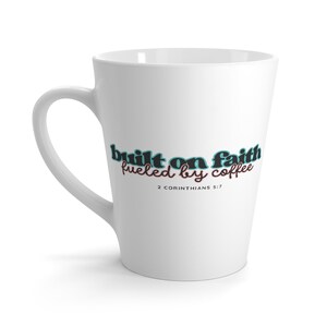 Może przedstawiać: Biały kubek do latte z zakrzywionym uchwytem. Kubek zawiera napis "built on faith fueled by coffee" w kolorze turkusowym i różowym, z wersetem biblijnym 2 Koryntian 5:7 poniżej.