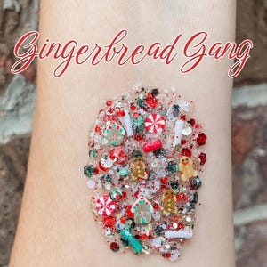 May include: A clear gel base filled with festive holiday-themed glitter and small charms, including miniature gingerbread people, houses, peppermint candies, and red, green, and white confetti. The text "Gingerbread Gang" is written in red script.