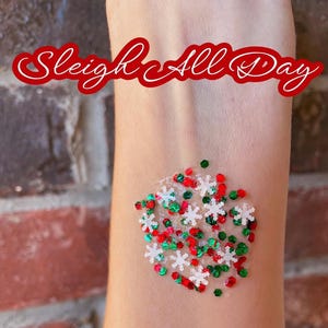 May include: A collection of loose cosmetic glitter featuring red, green, and white snowflake-shaped sequins. The text Sleigh All Day is written in a red cursive font above the glitter, which is applied to a patch of skin.