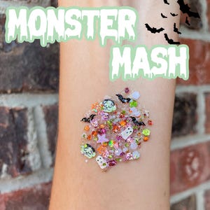 May include: A cluster of Halloween-themed glitter mix featuring small shapes of ghosts, bats, stars, and monster faces in orange, green, purple, and white. The text MONSTER MASH is displayed in a light green, dripping font above the glitter pile.