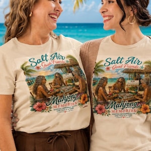 May include: A beige crew neck t-shirt featuring a colorful graphic print of three people playing mahjong on a beach. The text reads Salt Air Good Friends Mahjong Memories Keeping Memories Alive One Tile At A Time, surrounded by tropical flowers and palm trees.