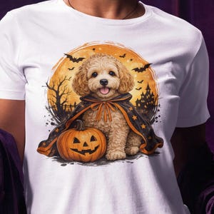 Puede incluir: Una camiseta blanca con el gráfico de un cachorro de pelo rizado que lleva una capa negra y naranja, sentado junto a una calabaza tallada. El fondo muestra una gran luna naranja, siluetas de murciélagos volando y un castillo oscuro.