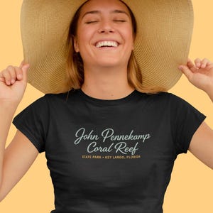 Op de afbeelding: Zwart t-shirt met de tekst "John Pennekamp Coral Reef State Park Key Largo, Florida" in een scriptlettertype. De persoon op de afbeelding draagt een grote strohoed en glimlacht.