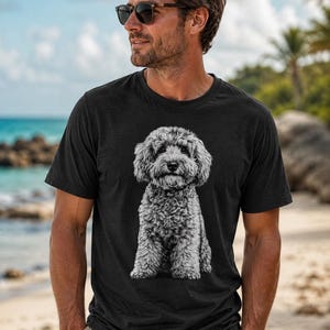 Op de afbeelding: Een zwart t-shirt met korte mouwen en een gedetailleerde, monochrome grafische print van een krulharige hond die rechtop zit. Het ontwerp is gecentreerd op de borst en toont de gestructureerde vacht en gelaatstrekken van de hond in grijstinten.