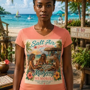 Op de afbeelding: Een perzikkleurig T-shirt met V-hals en een grafische print van drie mensen die mahjong spelen in een strandhut. De tekst luidt Salt Air Good Friends Mahjong MEMORIES KEEPING MEMORIES ALIVE, ONE TILE AT A TIME, omringd door tropische bloemen.