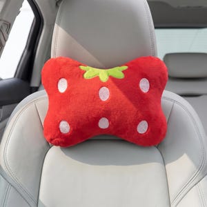May include: A red strawberry-shaped car headrest pillow with white polka dots and a green stem. The plush pillow is designed to fit on a car seat headrest. The pillow is made of soft, fuzzy material.