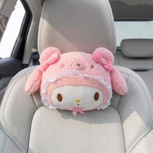 May include: A pink plush car headrest cover shaped like a cartoon character with a white face, brown eyes, and a small pink bow. The headrest has a fluffy texture, with pink bows on the ears and a lace trim around the face.