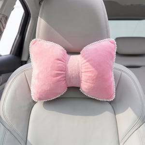 May include: A pink bow-shaped car headrest pillow with white pom-pom trim. The pillow is made of a soft, plush material and is positioned on a light gray car seat. The bow design adds a decorative touch to the car interior.