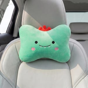 May include: A light green, plush car headrest shaped like a bone, with a cartoon dinosaur face. The headrest has black eyes, a smiling mouth, and pink cheeks. A red crest sits atop the headrest. The headrest is on a white car seat.