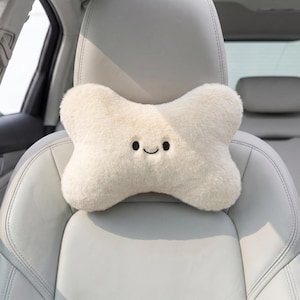 May include: A cream-colored, bone-shaped car headrest pillow with a smiling face. The plush pillow is positioned on a light gray car seat. The headrest is designed to provide comfort and support during car rides.