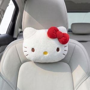 May include: White plush car headrest shaped like Hello Kitty, featuring brown eyes, a yellow nose, and a red bow. The headrest is designed to fit over a car seat headrest.