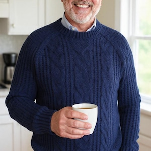 May include: A navy blue cable-knit sweater, light blue collared shirt, and blue jeans. A white mug is held in the hand. The background shows a kitchen with white cabinets and a window. The sweater has a textured pattern.